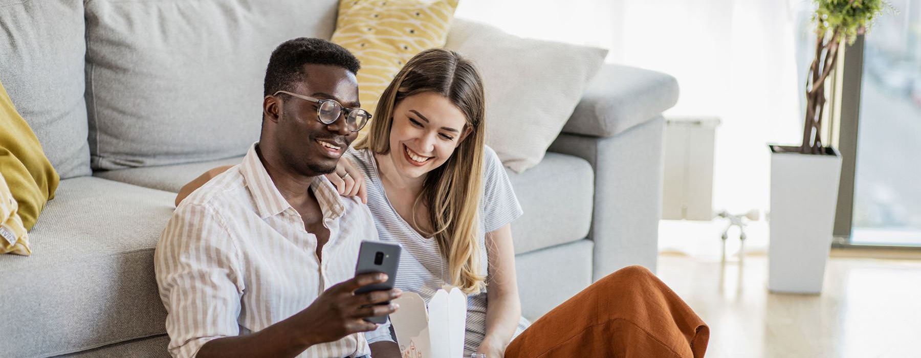 a man and woman with take out, sit against a couch on their living room floor and watch at their cell phone
